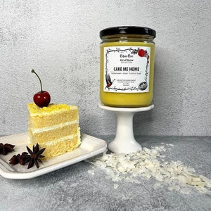 16 oz yellow cake candle on a pedastal. Beside the candle is a white plate with a piece of cake topped with a cherry. Candle label has a robin and a rose on it
