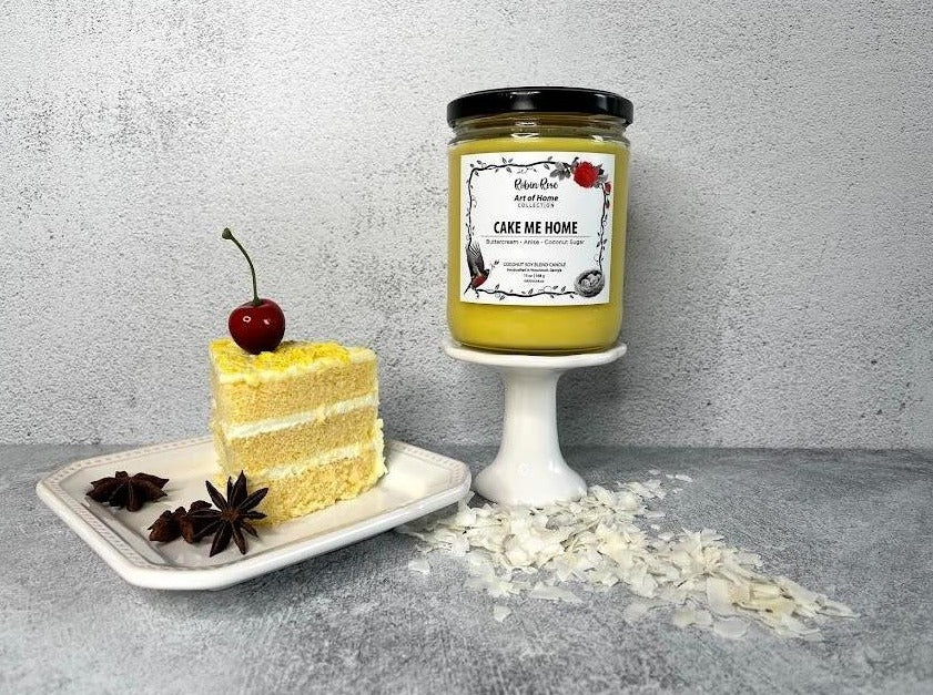16 oz yellow cake candle on a pedastal. Beside the candle is a white plate with a piece of cake topped with a cherry. Candle label has a robin and a rose on it